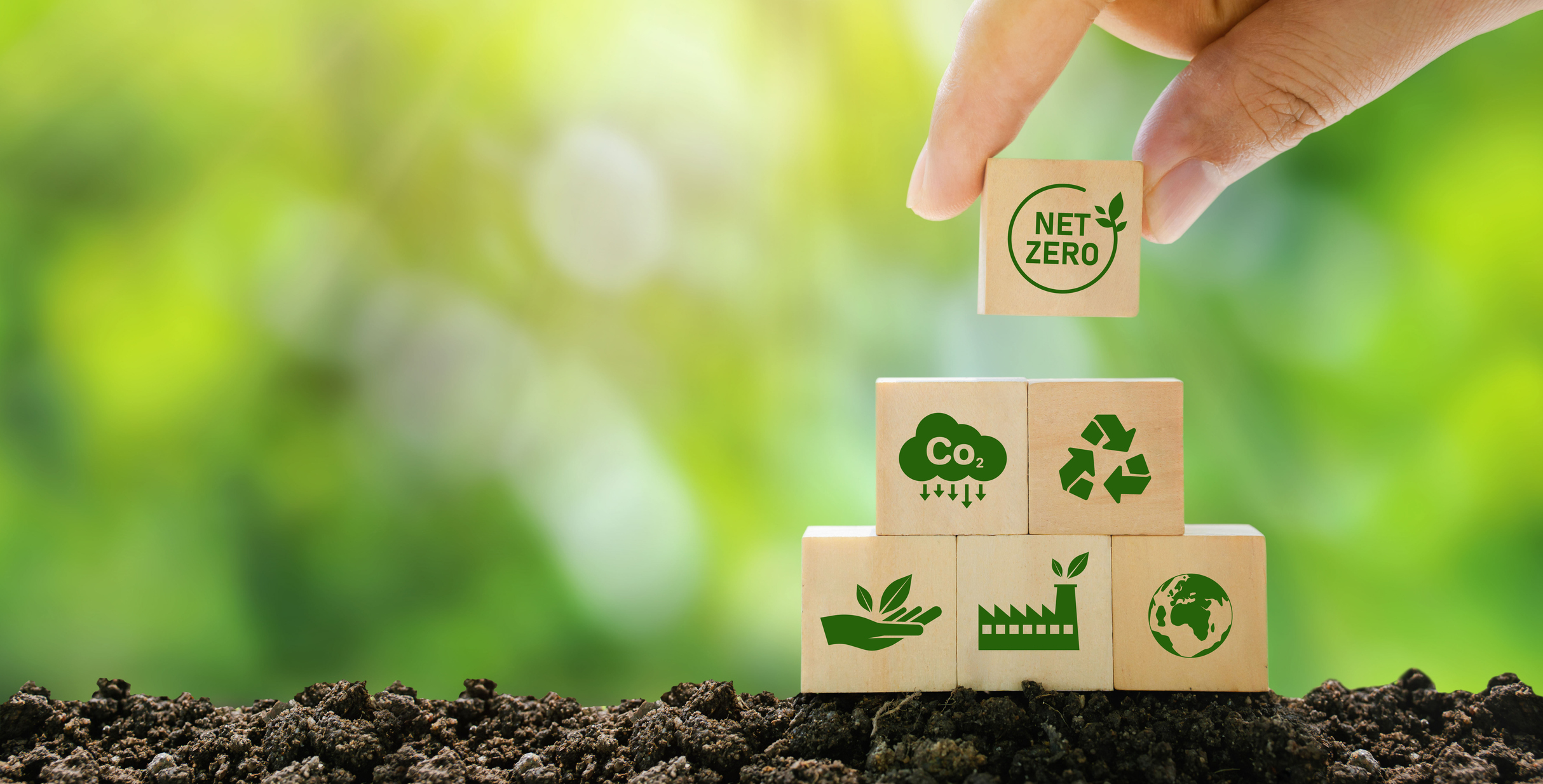 Hand placing the final wooden block labeled 'Net Zero' on top of a stack with other blocks displaying green energy and sustainability icons, against a blurred natural background.